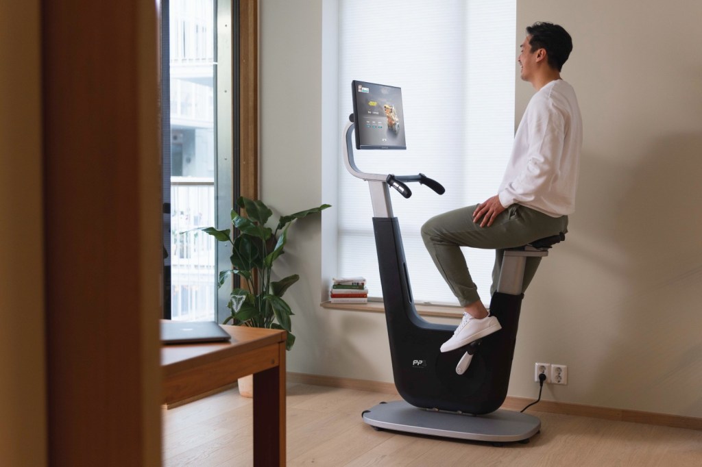 A man in a white sweatshirt, khaki joggers and white trainers sits on an exercise bike with a built-in screen. There is a plant in the corner and a laptop on the table in the foreground.