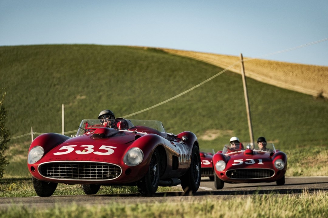 A still from the film Ferrari showing two red cars on a road with a green hill in the background